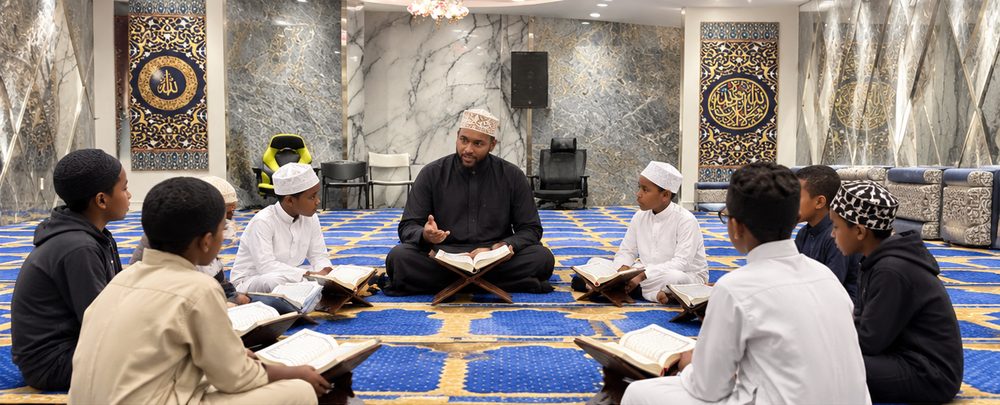 Boys in thawbs and kufis seated in a circle on the blue and gold prayer carpet of the Dar Al Nur hall, each with an open Qur’an on a wooden stand, facing their teacher.