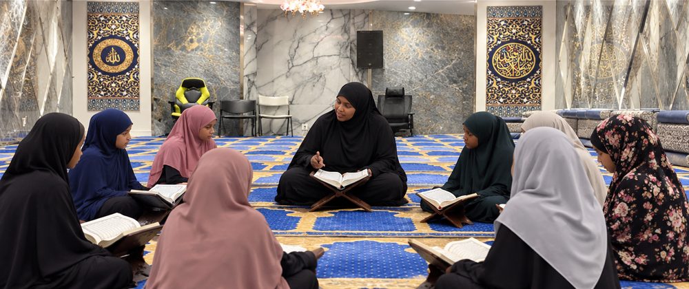 Girls in hijabs seated in a circle on the same prayer carpet, each with an open Qur’an on a wooden stand, facing their woman teacher.